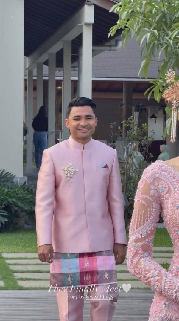 Seorang pria mengenakan setelan adat berwarna merah muda dengan kain bermotif kotak berdiri di area luar ruangan saat acara lamaran.