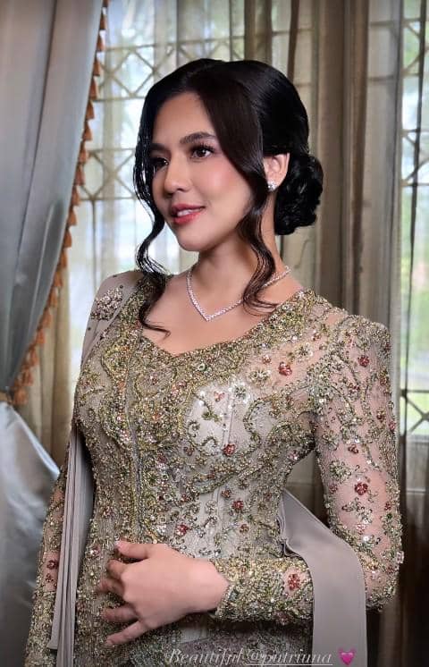 Seorang wanita mengenakan kebaya modern berwarna emas dengan hiasan payet dan rambut disanggul rapi di dalam ruangan berhias tirai.