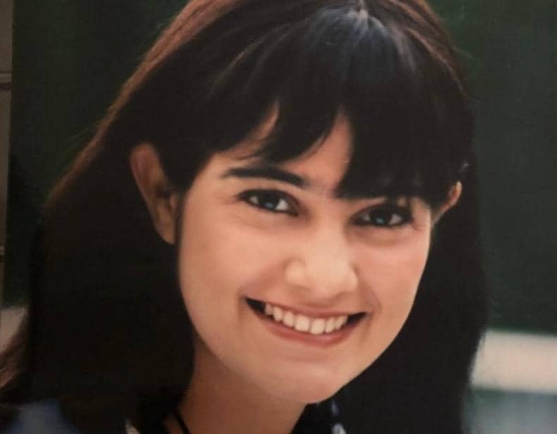 Seorang perempuan berambut cokelat gelap dengan poni tersenyum hangat ke arah kamera di latar luar ruangan.