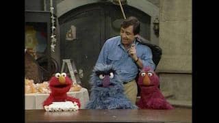 Seorang pria berdiri di belakang tiga karakter boneka berwarna merah, biru, dan ungu di set acara Sesame Street Classics: Season 1.