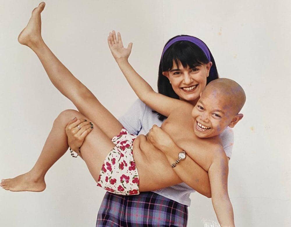 Seorang wanita tersenyum sambil menggendong anak laki-laki tanpa baju yang tertawa riang di latar belakang putih.
