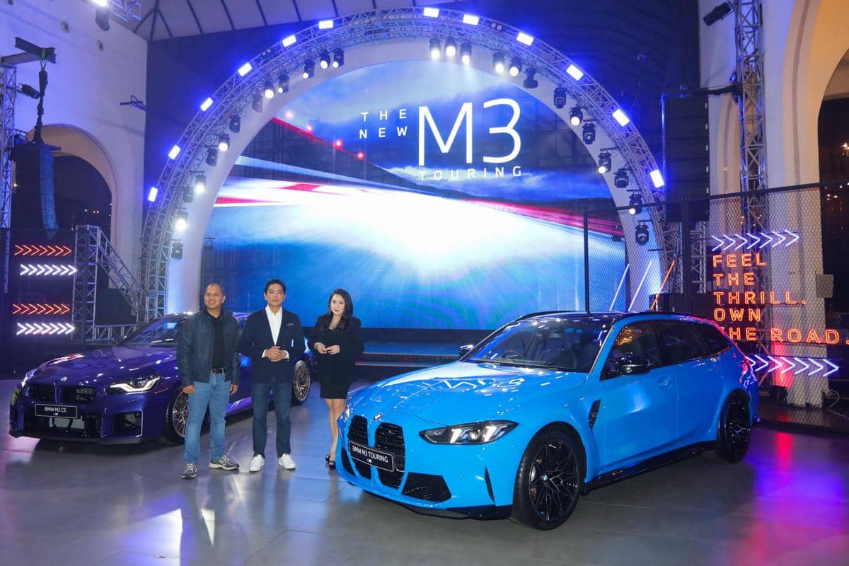 Tiga orang berdiri di samping dua mobil BMW M3 terbaru berwarna biru muda dan ungu dalam acara peluncuran di panggung dengan pencahayaan biru.