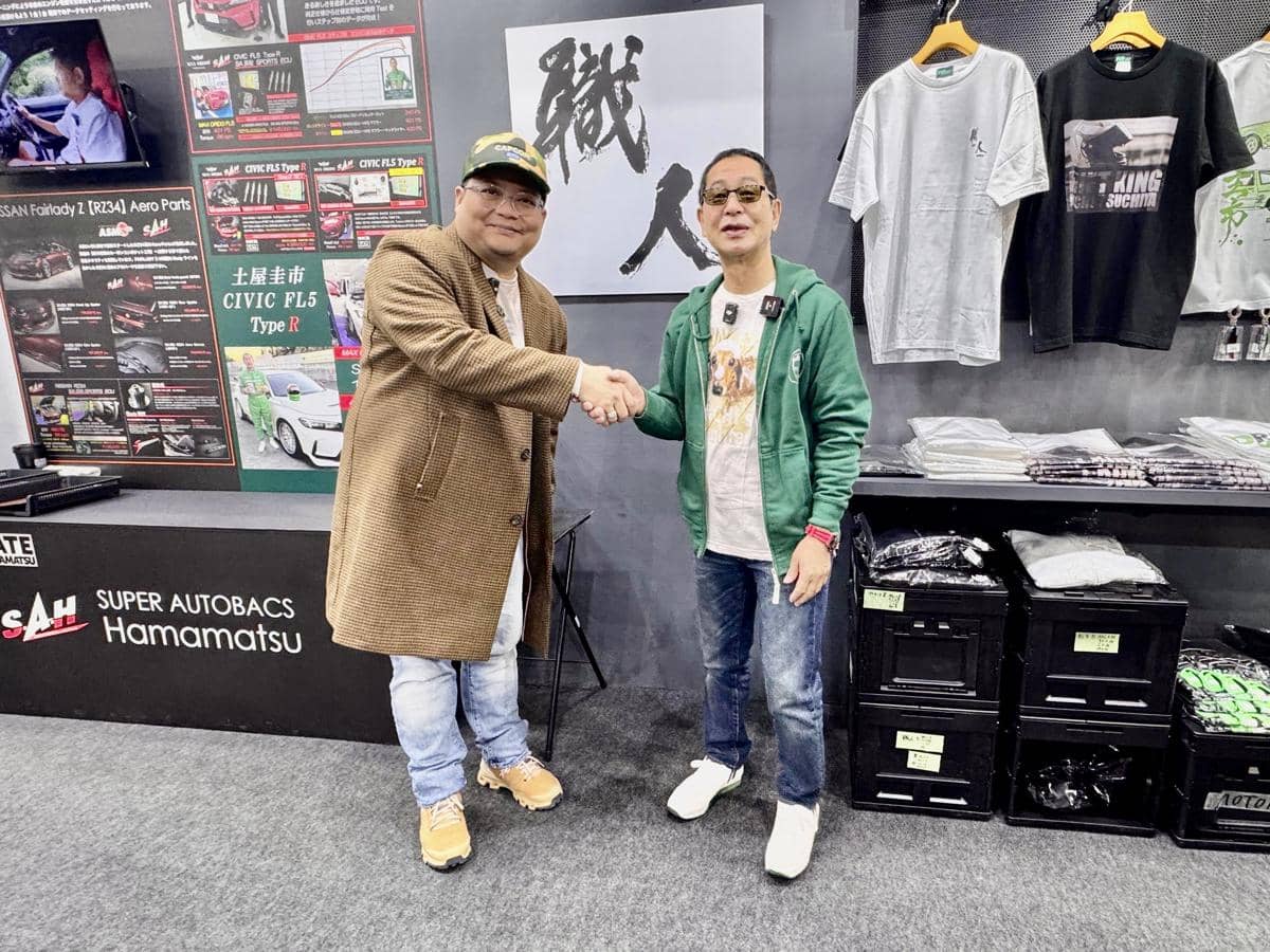 Dua pria berjabat tangan di stan Super Autobacs Hamamatsu dengan latar belakang poster mobil dan deretan kaus yang dipajang.