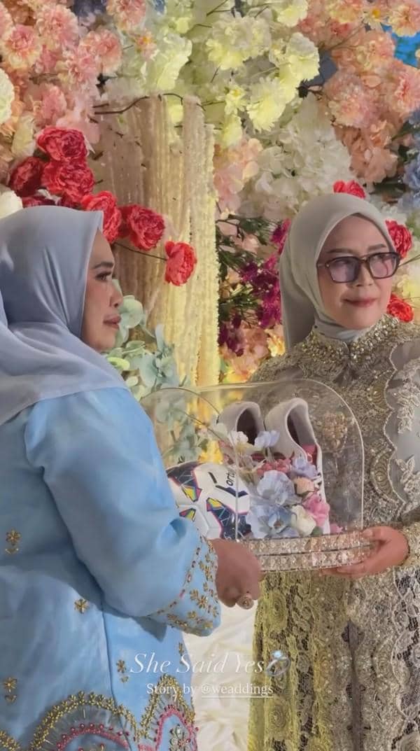 Dua wanita berhijab mengenakan busana tradisional elegan saling bertukar hantaran lamaran dengan latar dekorasi bunga warna-warni.