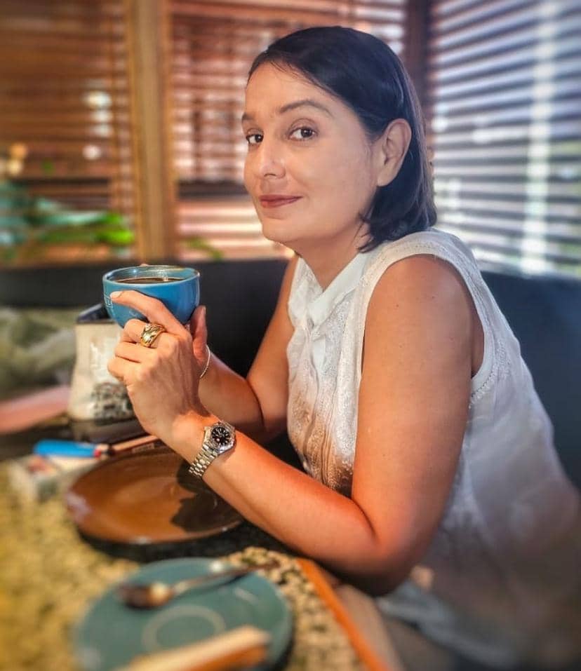 Seorang perempuan berambut pendek mengenakan blus putih duduk di meja kafe sambil memegang cangkir biru dan tersenyum ke arah kamera.