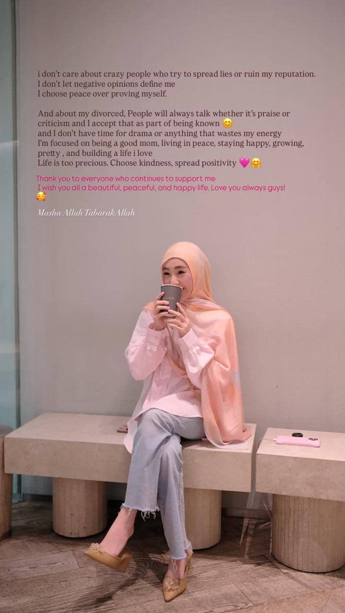 Seorang wanita berhijab berwarna peach duduk santai di bangku sambil memegang cangkir minuman, mengenakan atasan putih dan celana jeans.