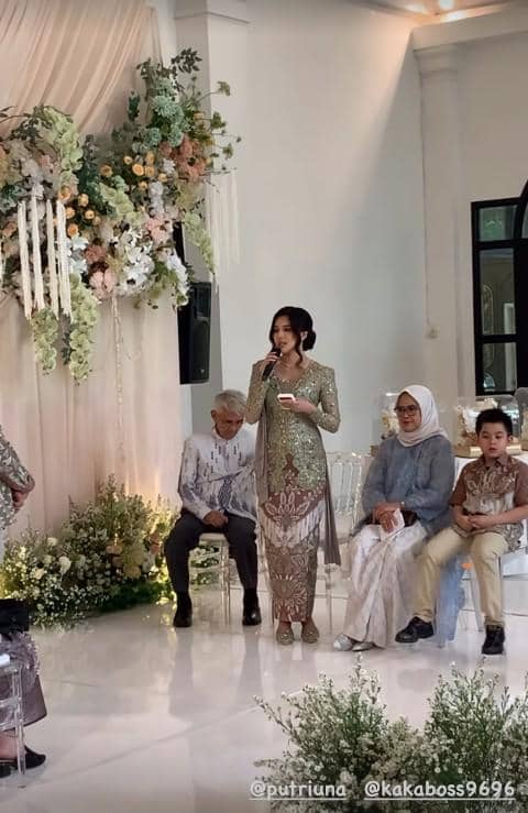 Seorang wanita mengenakan kebaya hijau berbicara dengan mikrofon di acara lamaran yang dihiasi bunga dan tirai putih elegan.