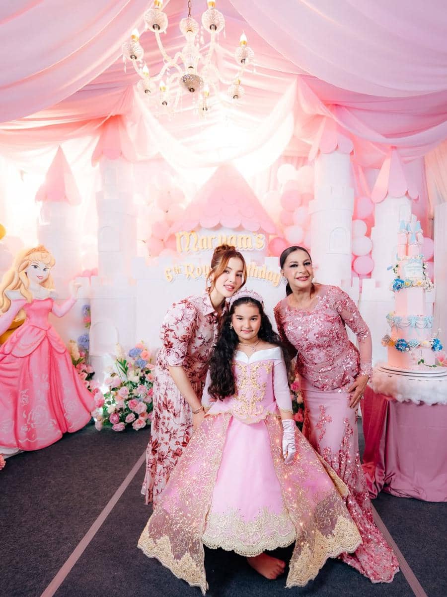 Seorang anak perempuan mengenakan gaun pesta berwarna pink berdiri di antara dua wanita dewasa di pesta ulang tahun bertema putri dengan dekorasi balon dan kue.