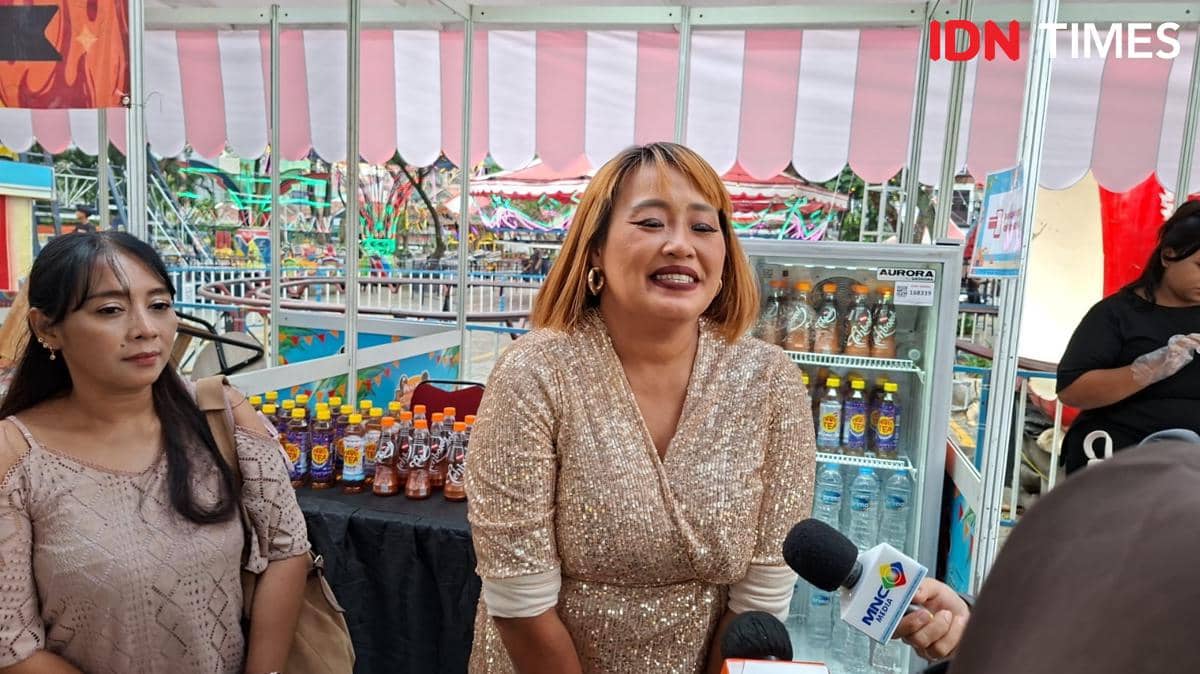 Seorang perempuan mengenakan gaun berkilau sedang tersenyum di stan minuman dan makanan di TangCity Mall, dikelilingi wartawan.