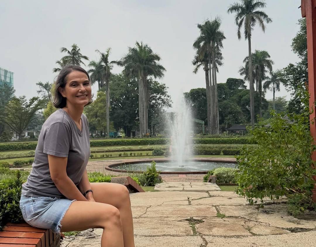 Seorang wanita tersenyum duduk di tepi taman dengan air mancur di latar belakang dan deretan pohon palem yang menjulang tinggi.
