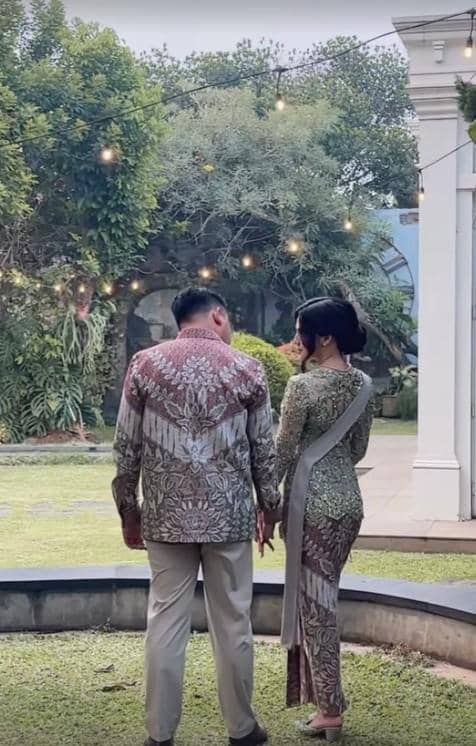 Sepasang pria dan wanita mengenakan busana batik tradisional berdiri berhadapan di taman dengan lampu hias menggantung di atas mereka.