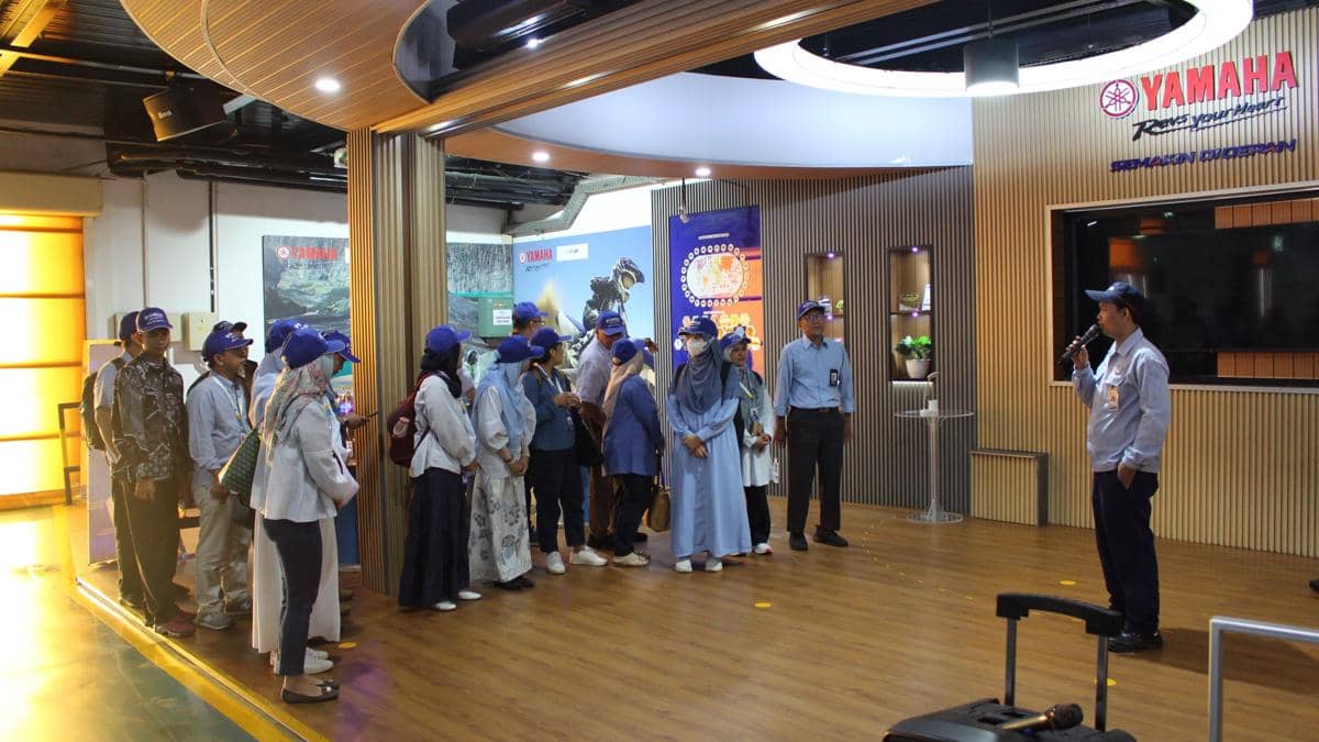 Sekelompok orang mengenakan topi biru mendengarkan penjelasan dari seorang pembicara di area pameran Yamaha dengan latar logo perusahaan.