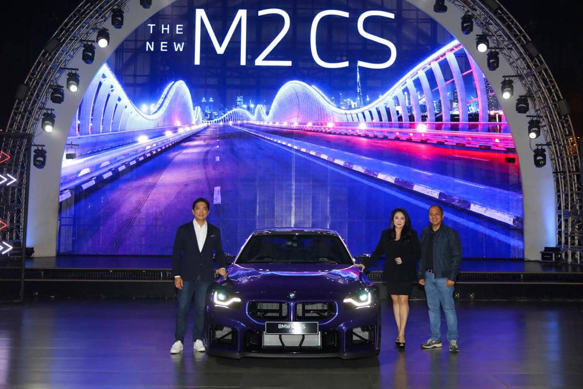 Tiga orang berdiri di samping mobil BMW M2 CS berwarna ungu saat acara peluncuran dengan latar belakang layar besar bertema jalan malam.