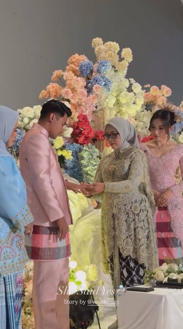 Prosesi lamaran sepasang calon pengantin berpakaian adat modern dengan latar bunga warna-warni dan keluarga yang mendampingi.