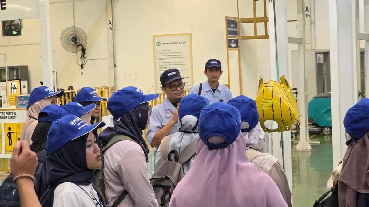 Seorang pemandu memberikan penjelasan kepada sekelompok peserta yang mengenakan topi biru di dalam area pabrik atau fasilitas industri.