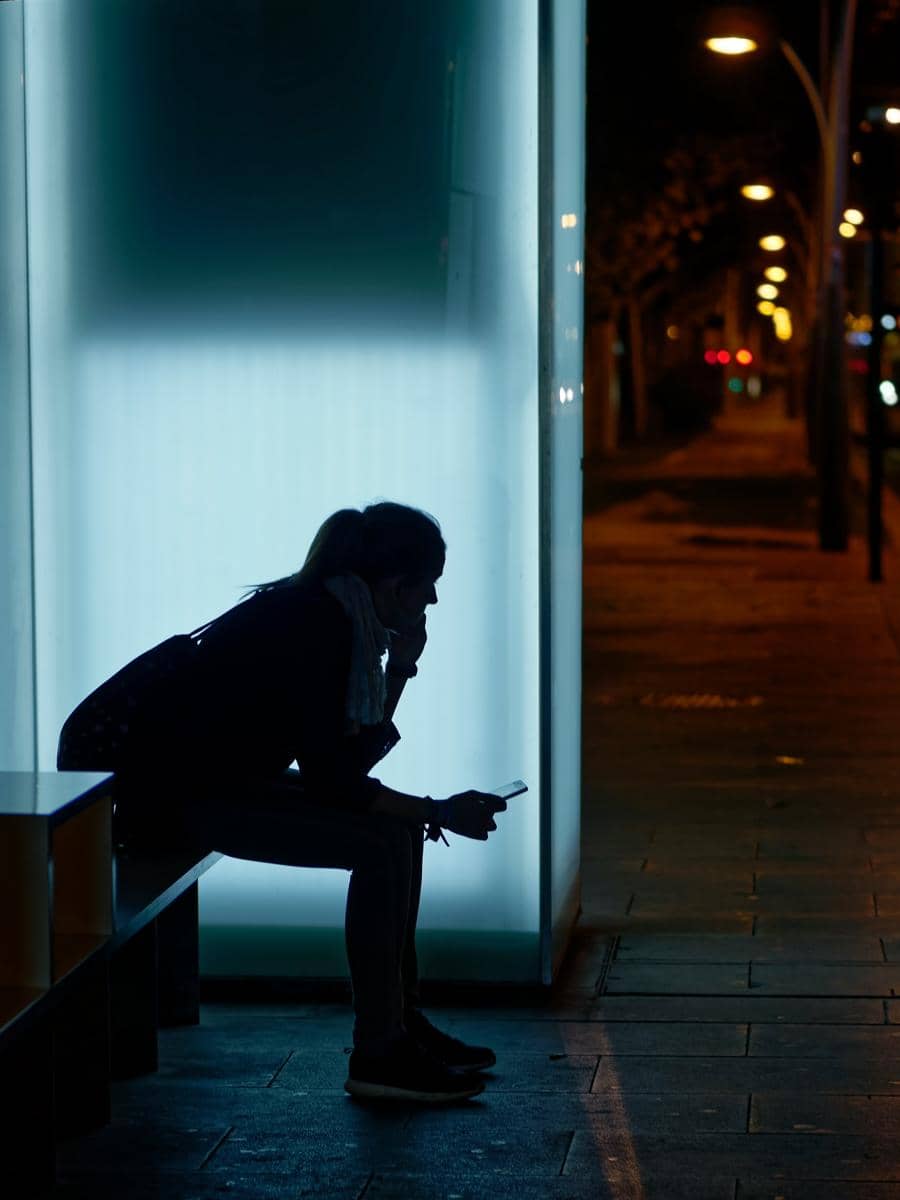 Siluet seorang perempuan duduk sendirian di halte malam hari sambil memegang HP dengan ekspresi lelah.