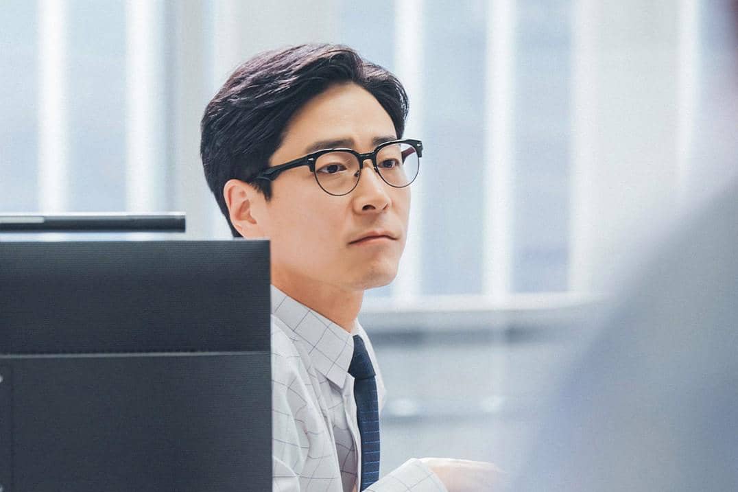 Seorang pria berkacamata mengenakan kemeja kotak dan dasi duduk di meja kantor dengan komputer di depannya dalam adegan drakor See You at Work Tomorrow.