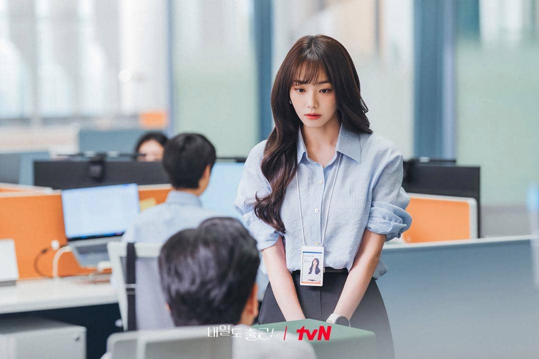 Seorang wanita mengenakan kemeja biru muda berdiri di depan meja kantor sambil berbicara dengan rekan kerja di ruang kerja modern.