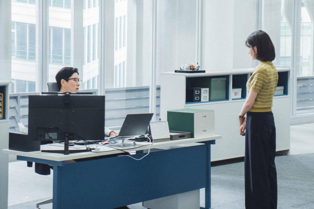 Dua orang sedang berbicara di kantor modern dengan meja kerja dan komputer, diambil dari drama Korea See You at Work Tomorrow.