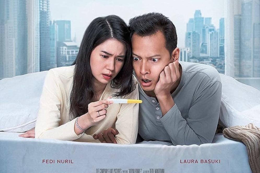 Sepasang suami istri di atas tempat tidur menatap alat tes kehamilan dengan ekspresi terkejut dan penasaran di latar kota modern.