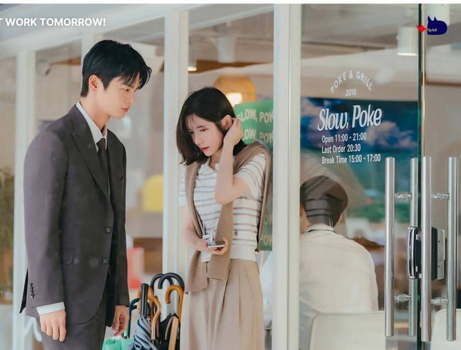 Seorang pria berjas dan seorang wanita dengan pakaian kasual berdiri di depan restoran Slow Poke dalam cuplikan drama See You at Work Tomorrow.