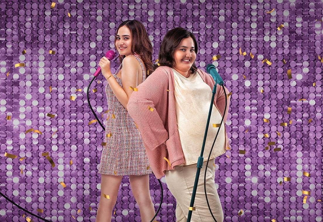 Dua wanita berdiri bersebelahan sambil memegang mikrofon di depan latar ungu berkilau dengan pita emas bertebaran.