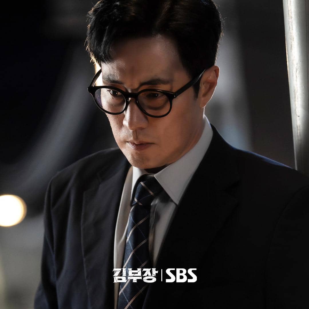 Seorang pria mengenakan setelan jas hitam dan kacamata tampak serius dalam cuplikan drama Agent Kim Reactivated produksi SBS.