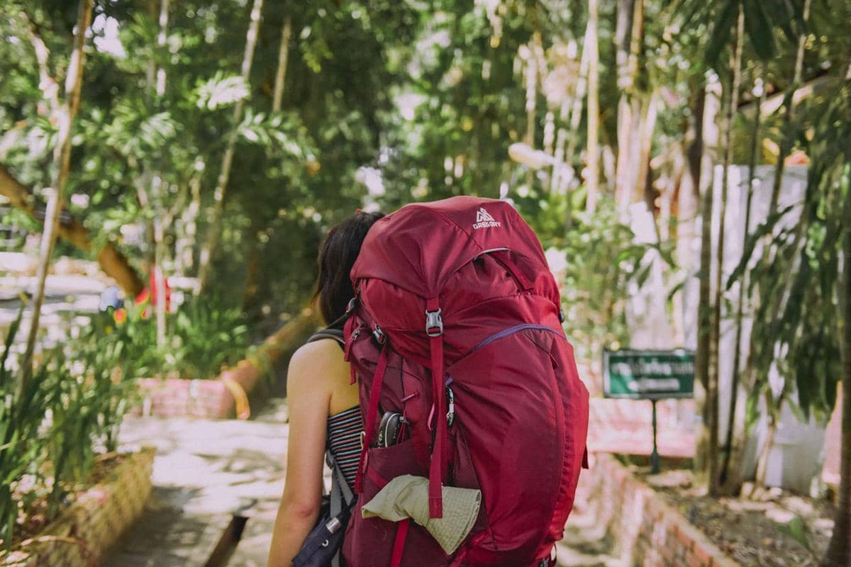 Seorang perempuan berjalan di jalur taman dengan membawa ransel besar berwarna merah di punggungnya di bawah pepohonan rindang.