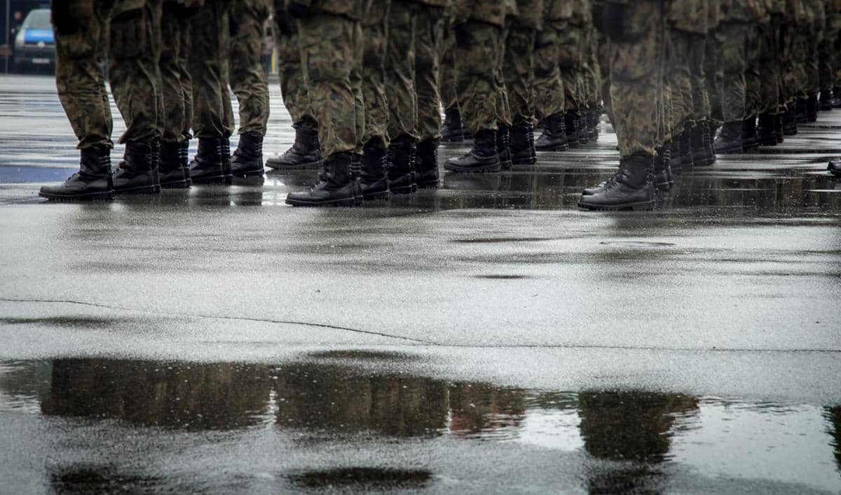 Barisan tentara mengenakan seragam kamuflase berdiri di atas permukaan jalan basah saat latihan militer di lapangan terbuka.