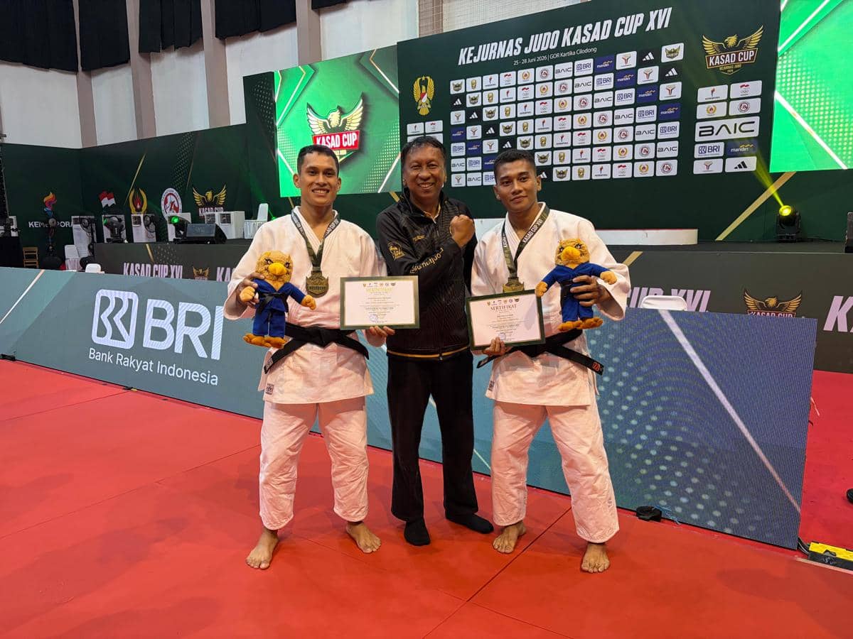 Tiga pria berpose di arena judo, dua di antaranya mengenakan gi putih sambil memegang sertifikat dan boneka usai meraih medali emas.