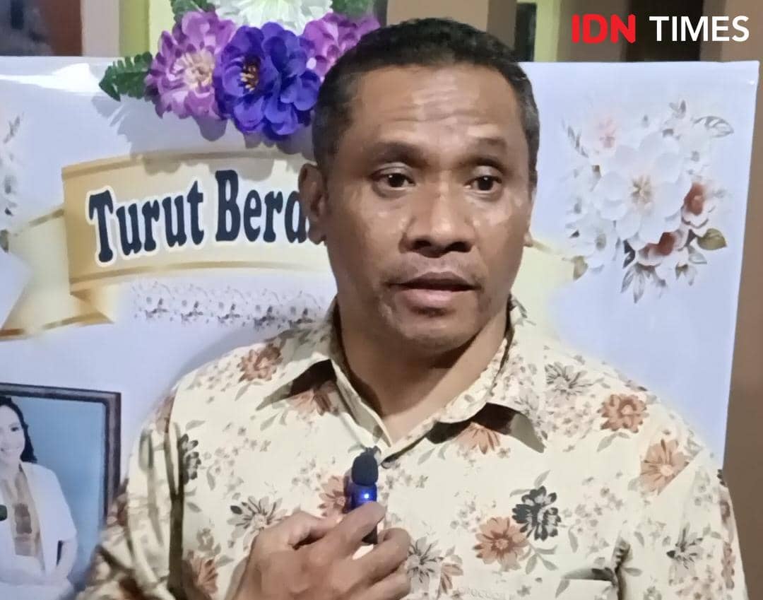 Seorang pria mengenakan kemeja bermotif bunga sedang berbicara di depan poster bertuliskan ucapan belasungkawa dengan hiasan bunga.
