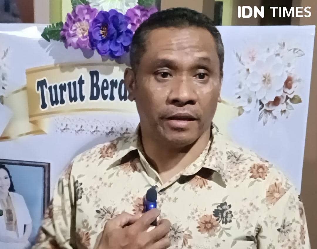 Seorang pria mengenakan kemeja bermotif bunga sedang berbicara di depan poster bertuliskan ucapan belasungkawa dengan hiasan bunga.