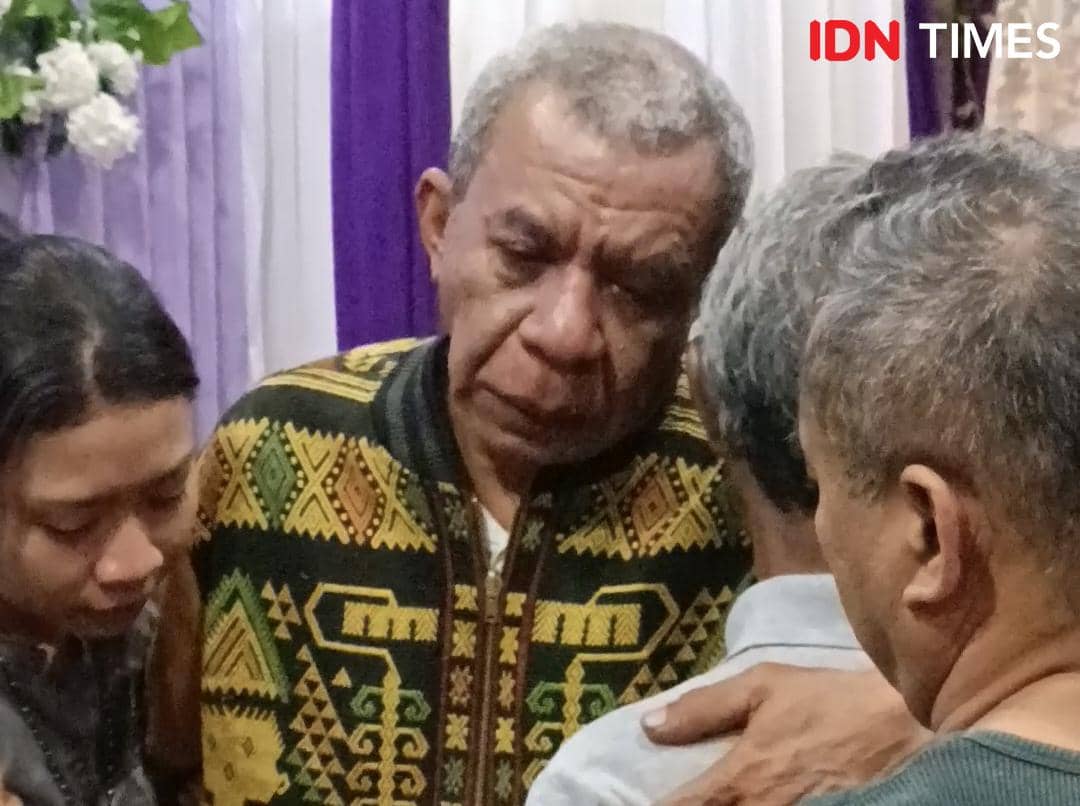 Seorang pria paruh baya mengenakan pakaian bermotif etnik sedang menangis dan dipeluk oleh beberapa orang di sekitarnya.