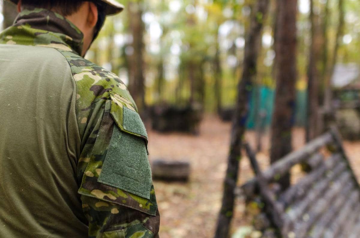 Seorang pria mengenakan seragam kamuflase berdiri di area hutan dengan latar pepohonan dan perlengkapan latihan militer.