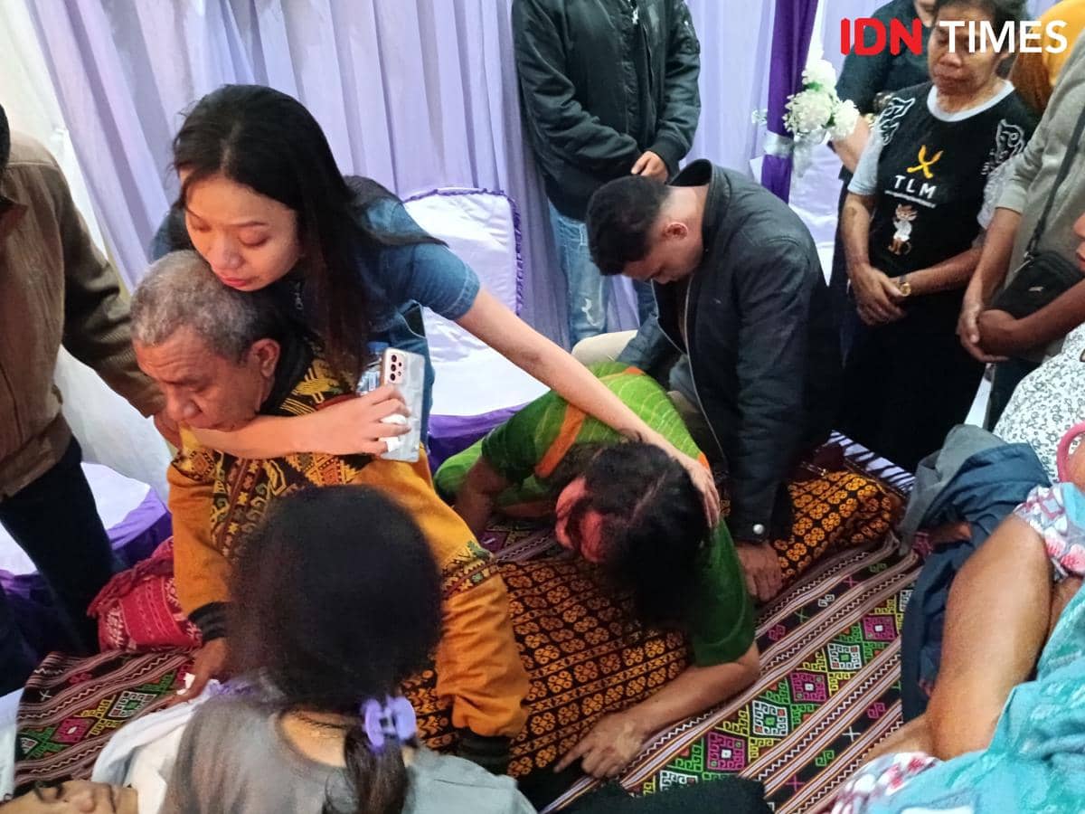 Keluarga berduka memeluk dan menangis di samping jasad seorang perempuan yang terbaring di kain bermotif tradisional saat prosesi perpisahan.
