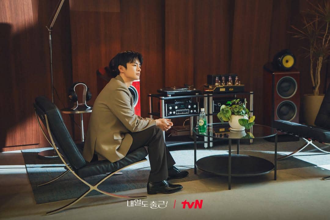 Seorang pria mengenakan setelan jas duduk di kursi modern di ruangan bergaya retro dengan peralatan audio dan tanaman hias di meja kaca.