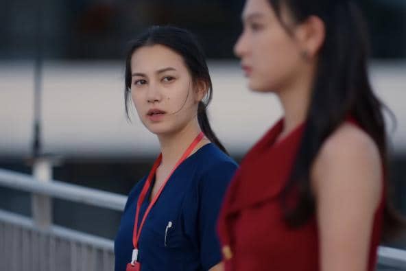 Seorang perempuan mengenakan seragam medis biru dengan lanyard merah menatap perempuan lain berbaju merah di area luar ruangan.