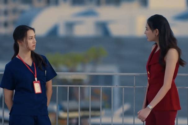 Dua wanita berdiri dan berbicara di luar ruangan, satu mengenakan seragam medis biru dengan ID card merah, dan satu mengenakan setelan merah tanpa lengan.