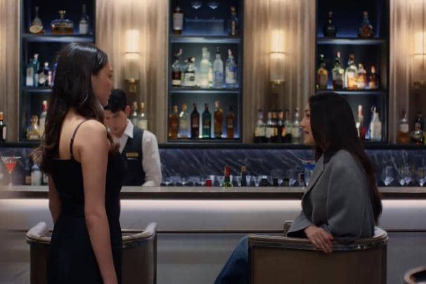 Dua wanita berbicara di sebuah bar modern dengan latar rak berisi botol minuman, sementara seorang bartender bekerja di belakang mereka.