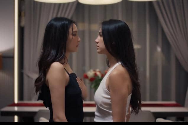 Dua wanita berdiri saling berhadapan di dalam ruangan dengan pencahayaan hangat, satu mengenakan gaun hitam dan satu mengenakan atasan putih.