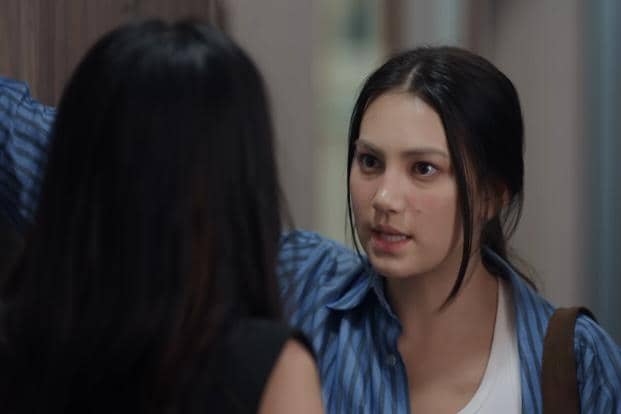 Seorang wanita berambut hitam mengenakan kemeja biru berbicara serius dengan wanita lain di sebuah ruangan sempit.