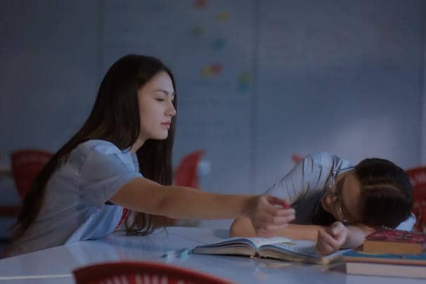 Dua siswi berseragam duduk di meja belajar, satu tertidur di atas buku sementara yang lain meraih tangannya di ruang kelas redup.