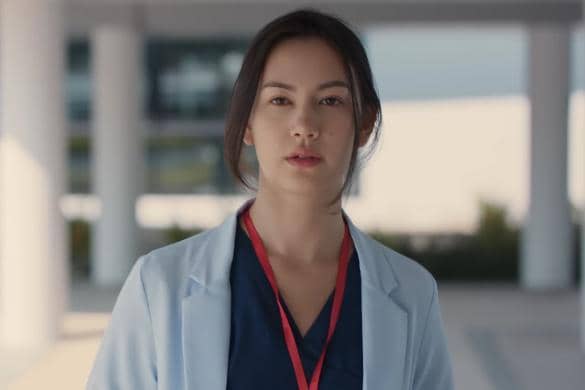 Seorang perempuan mengenakan jas dokter biru muda dan lanyard merah berdiri di luar gedung dalam cuplikan drama Did You Know.