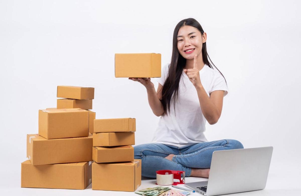 Seorang wanita duduk di lantai sambil memegang kotak paket dengan laptop di depannya dan beberapa kotak kardus di sekelilingnya.
