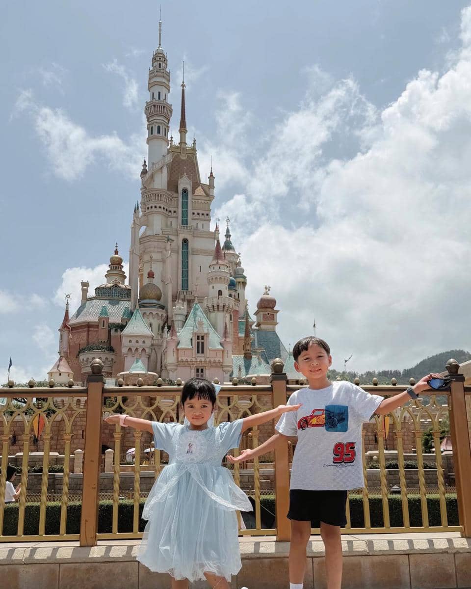 Dua anak kecil berpose ceria dengan tangan terbuka di depan kastel ikonik Disneyland Hong Kong di bawah langit cerah.