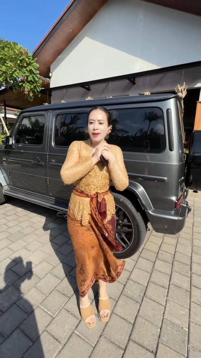 Seorang wanita mengenakan kebaya berwarna emas dan kain batik cokelat berpose di depan mobil SUV hitam di halaman rumah.