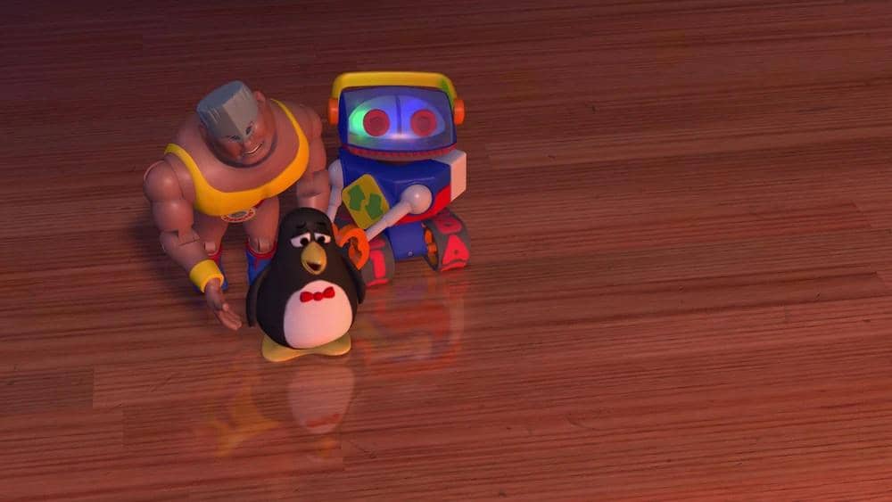 Tiga mainan karakter animasi terdiri dari robot berwarna cerah, penguin kecil, dan figur berotot berdiri di lantai kayu.