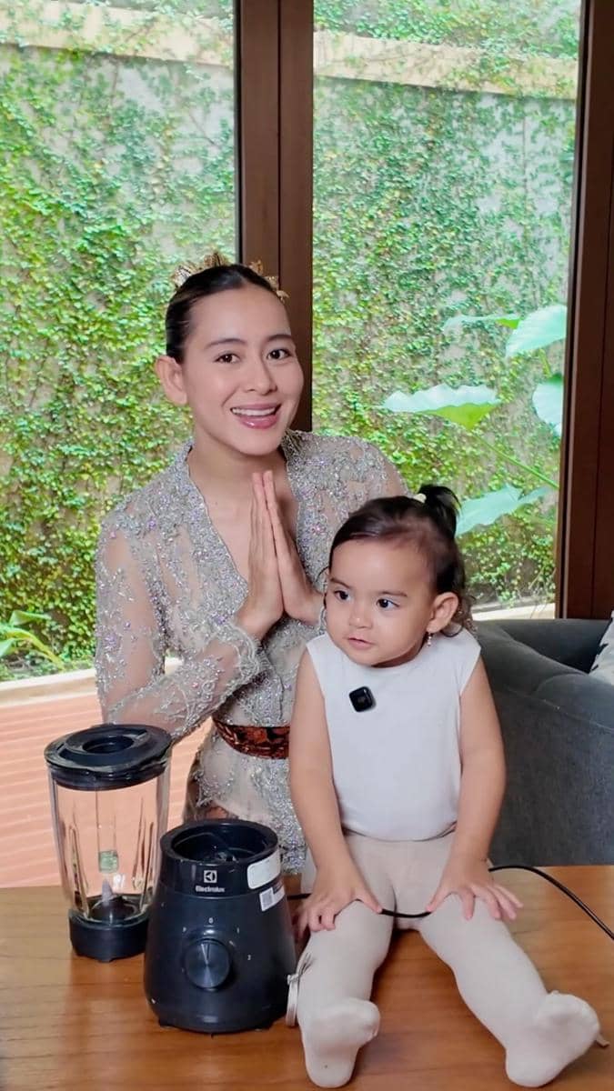 Seorang wanita mengenakan kebaya berwarna lembut berpose dengan tangan menyatu di samping anak kecil yang duduk di meja kayu di dalam rumah.