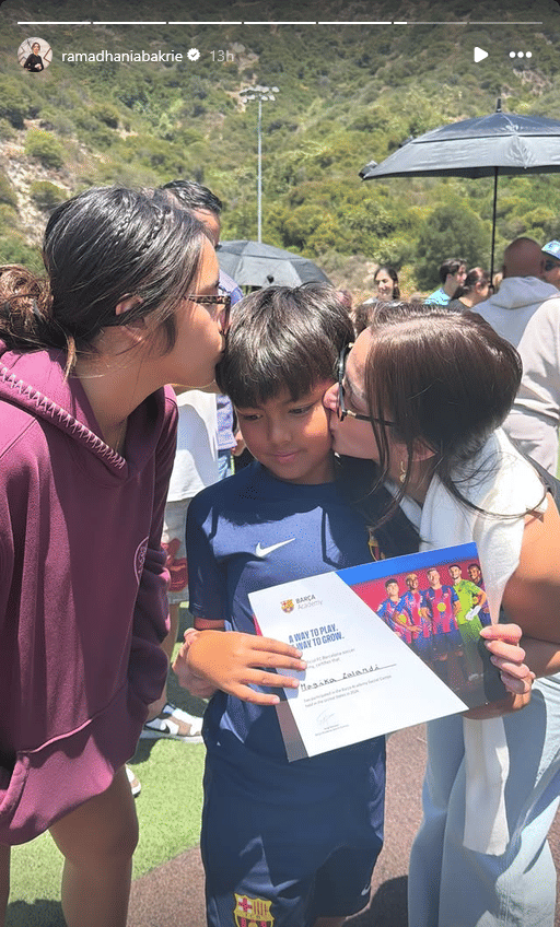 Seorang anak mengenakan seragam sepak bola Barcelona Academy memegang sertifikat sambil dicium di pipi oleh dua wanita di area luar ruangan.