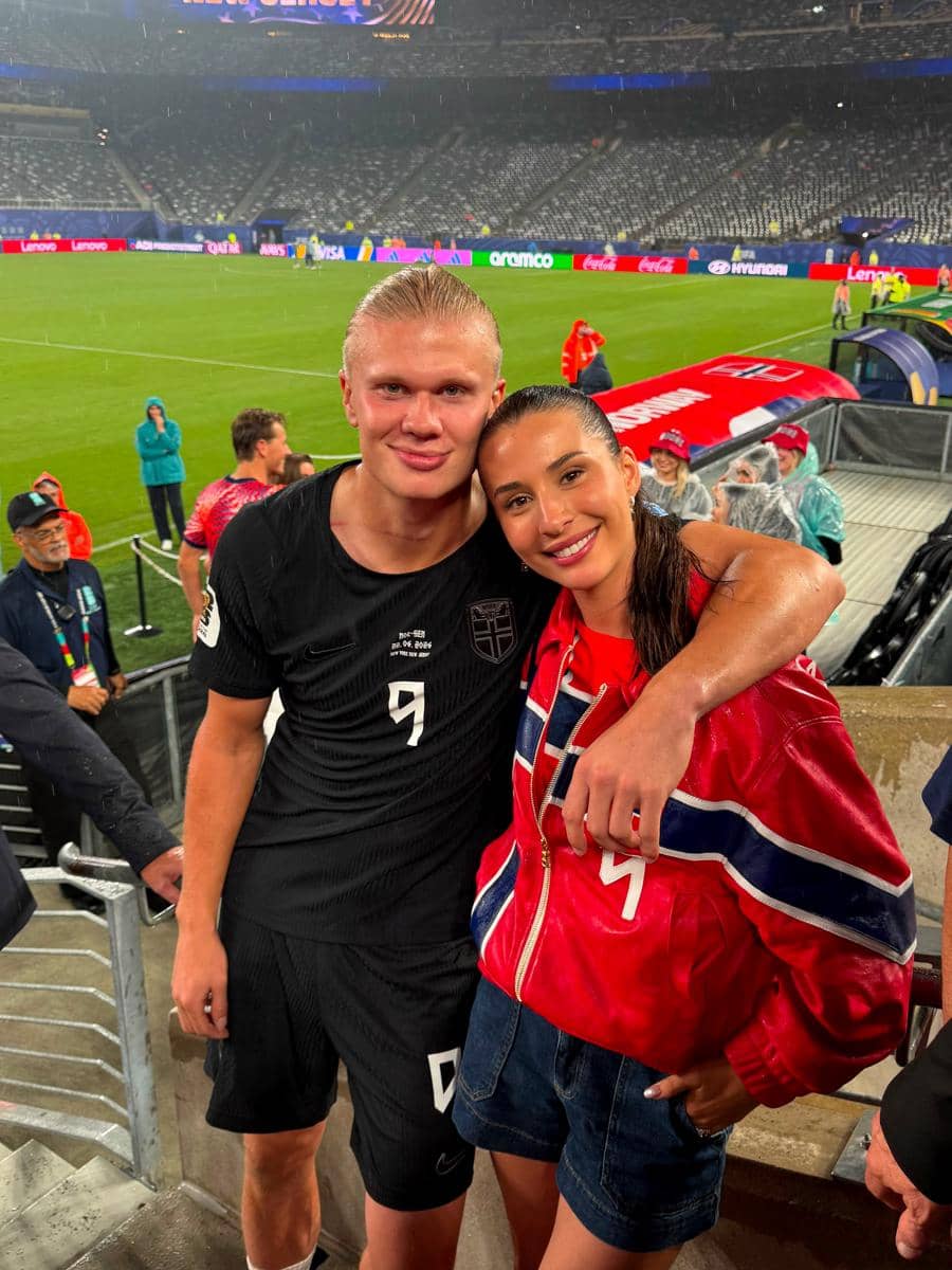 Seorang pemain sepak bola mengenakan seragam hitam berpose bersama seorang wanita berjaket merah di stadion seusai pertandingan.
